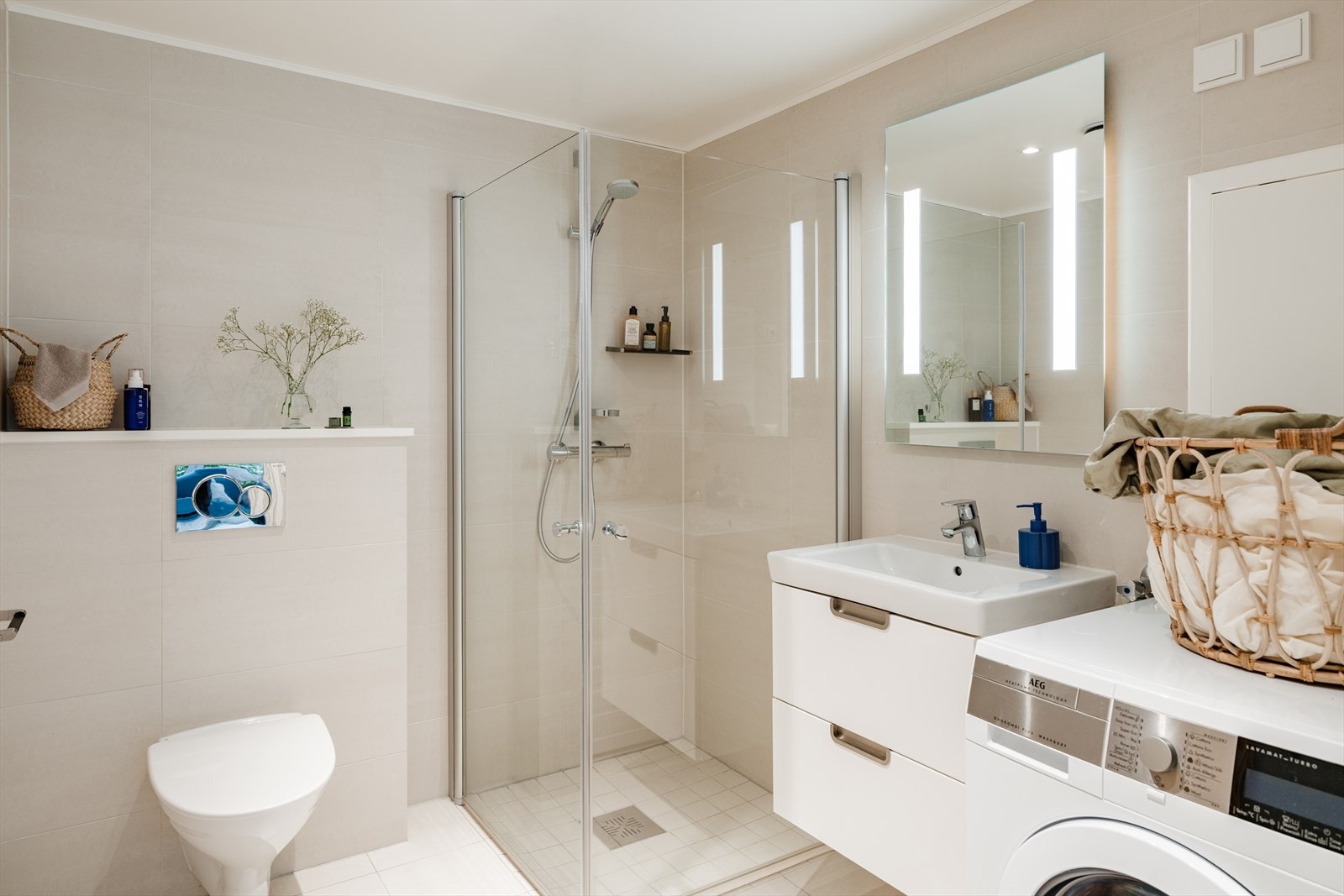 Badet er av god størrelse og er utstyrt med vegghengt toalett, dusjhjørne med glassdører, servant med underskap, speil med integrert belysning samt opplegg for vaskemaskin Galleribilde
