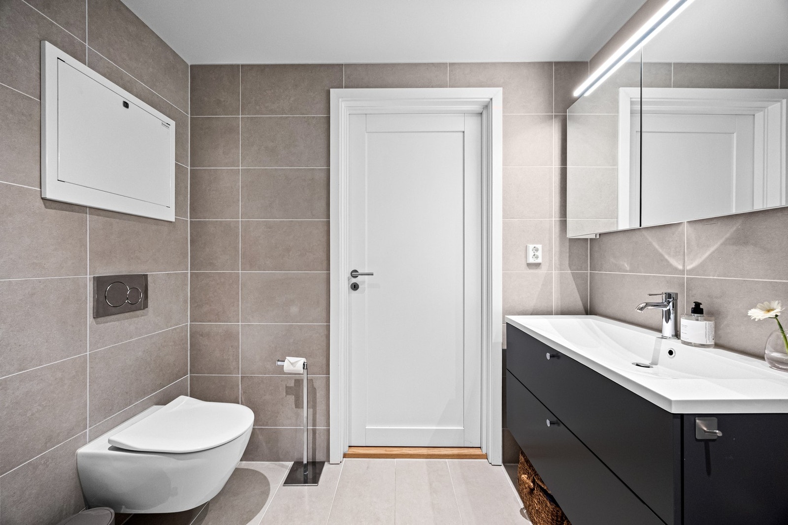 Badet er utstyrt med en baderomsinnredning fra Sigdal med glatte, fabrikklakkerte fronter og heldekkende servant, samt et speilskap med integrert lys. Det er opplegg for vaskemaskin, veggmontert toalett og et dusjhjørne med glassdører. Galleribilde