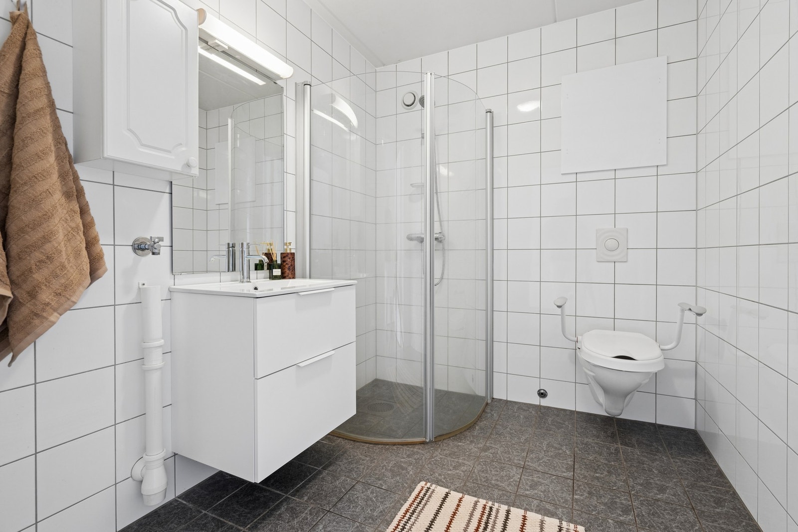 Badet er helfliset og har elektrisk gulvvarme. Rommet er utstyrt med vegghengt toalett, dusjhjørne med glassdører og opplegg for vaskemaskin. Galleribilde