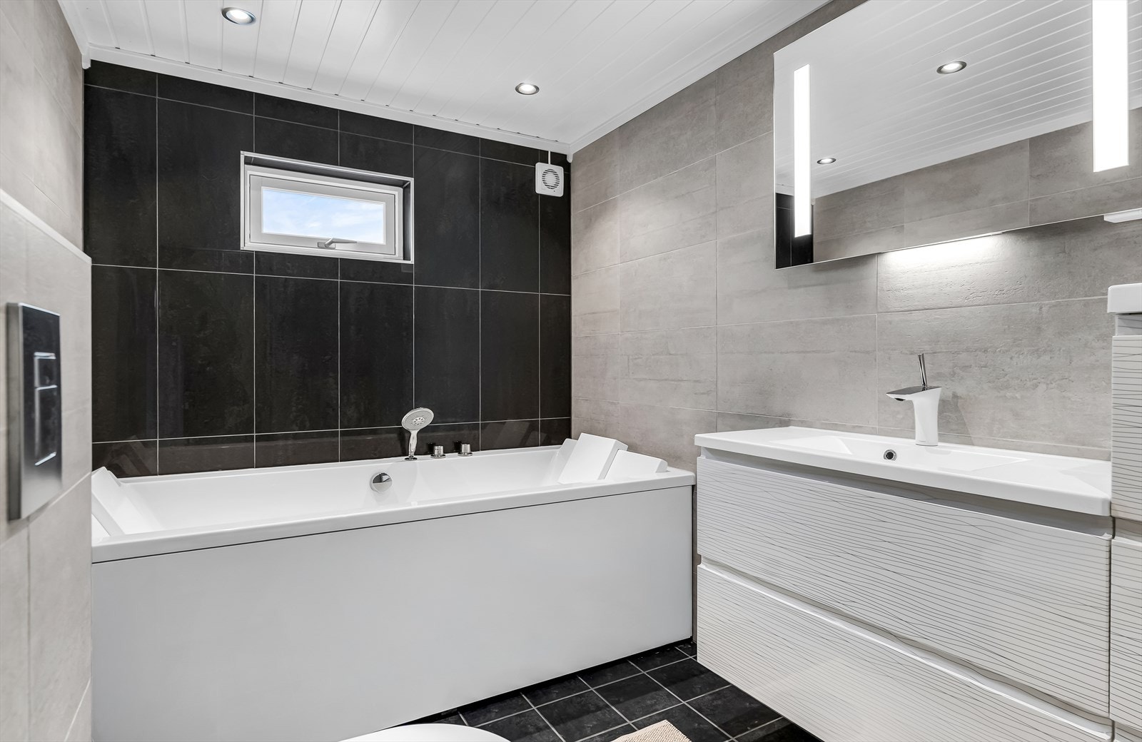 Badet er utstyrt med servantinnredning med servanttopp, speil med integrert belysning, romslig badekar, vegghengt toalett og dusjhjørne med dusjopplegg på vegg. Galleribilde