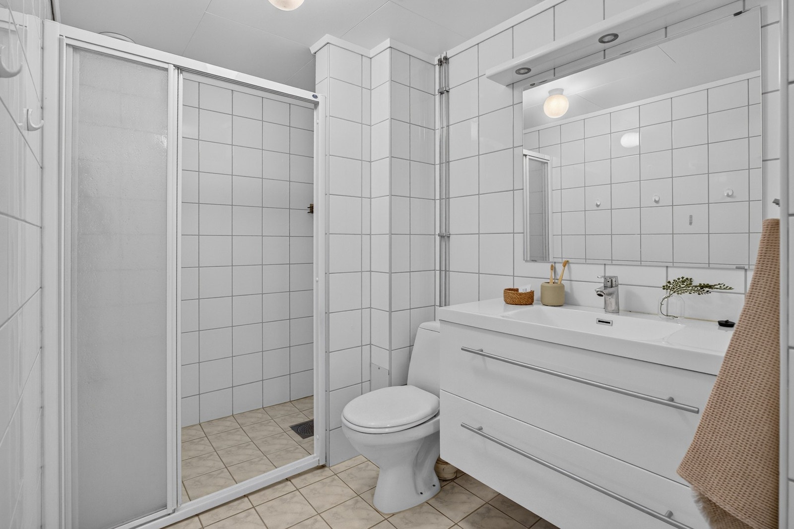 Badet er innredet med hvite veggfliser og lysegrå fliser på gulvet. Varme er lagt i gulvet, og badet har toalett og et dusjhjørne med skyvedør Galleribilde
