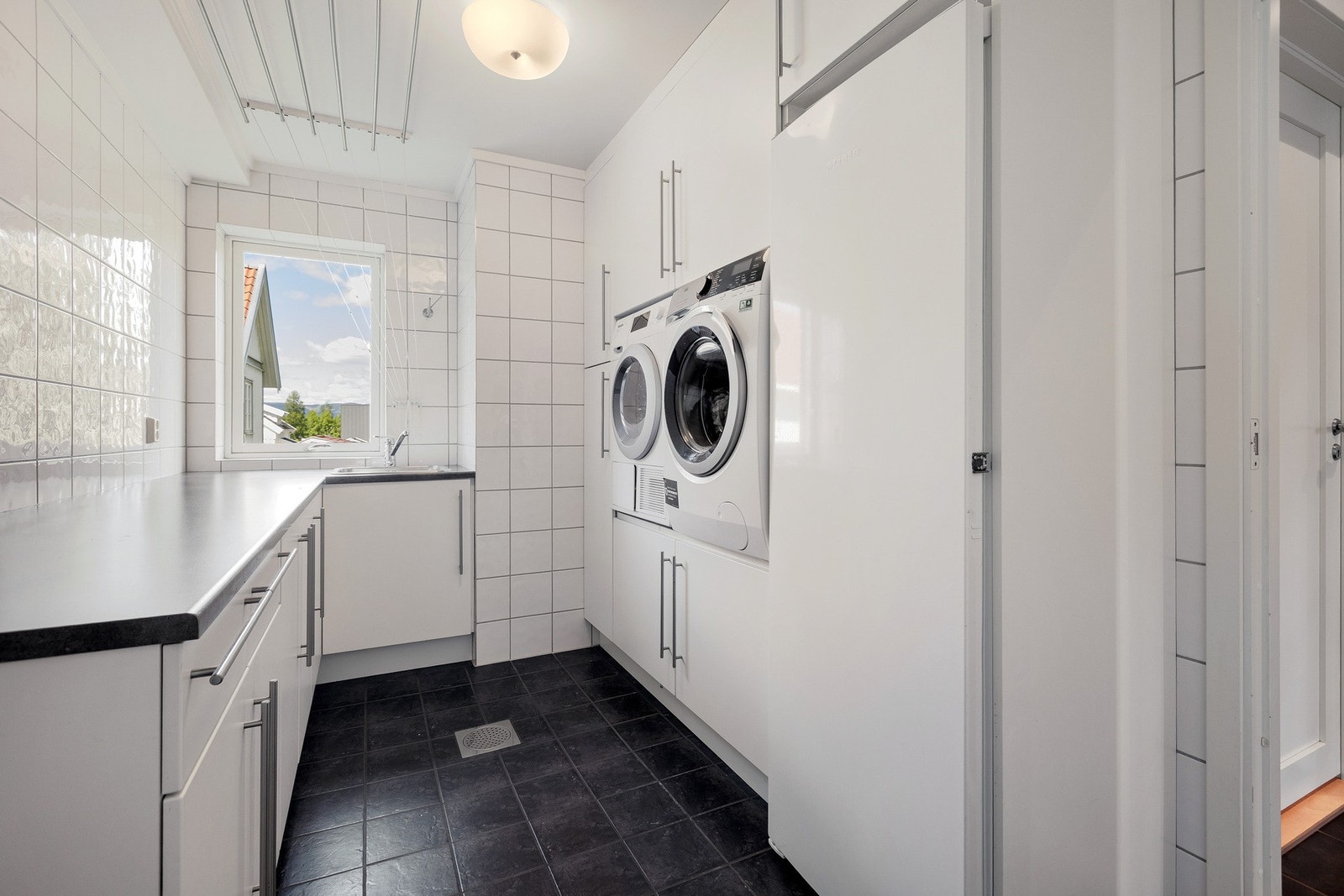 Romslig og praktisk flislagt vaskerom med opplegg for vaskemaskin og tørketrommel plassert i arbeidshøyde i innredning. Galleribilde