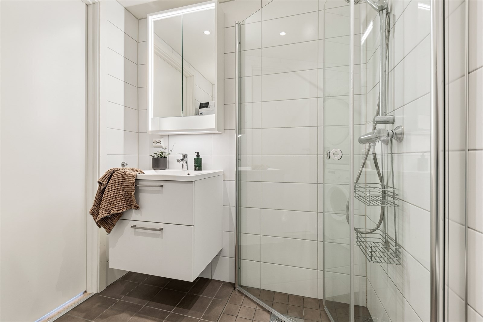 Badet har flislagte overflater, vegghengt toalett og dusjhjørne med innfellbare glassdører. Galleribilde