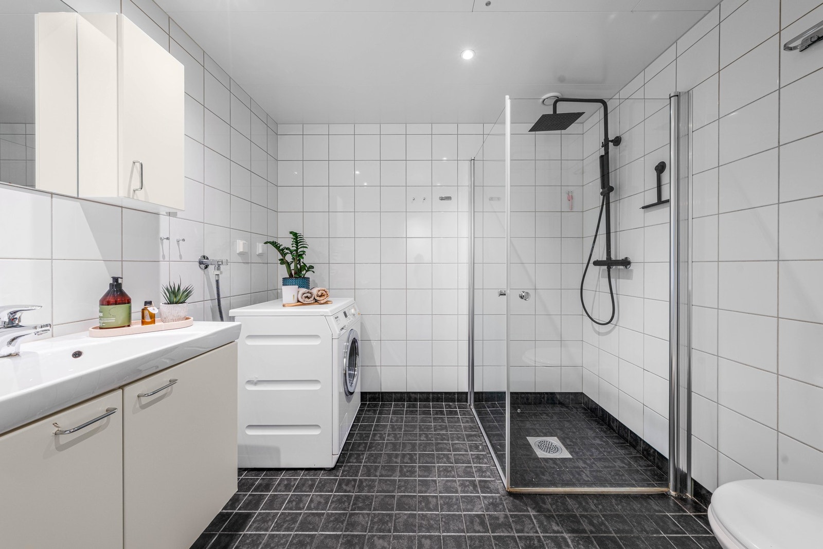 Baderommet er utstyrt med lys baderomsinnredning med heldekkende servant, dusjhjørne med innfellbare glassdører og både hånd- og regnfallsdusj, samt vegghengt toalett. I hjørnet er det plass og opplegg for vaskemaskin. Galleribilde
