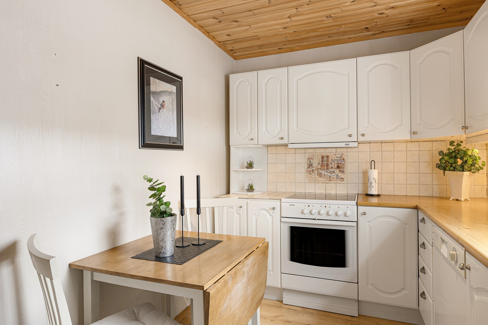 Over benken er det belysning og beige fliser, og ventilatoren over kokesonen er fra 2025. Deler av benken er byttet ut med samme type fliser som mellom overskap og benk. Kjøkkenet har opplegg for oppvaskmaskin og avsatt plass til komfyr. Galleribilde