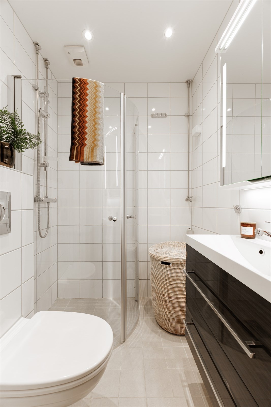 Badet fremstår lyst og tidløst med lyse fliser og en praktisk planløsning. Baderomsinnredningen består av benkeskap med glatte fronter samt veggskap med speilfronter som gir god oppbevaringsplass. Galleribilde