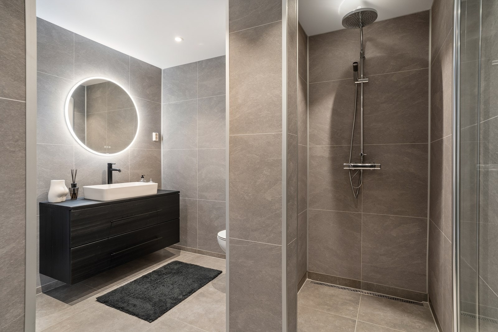 Badet er utstyrt med servantinnredning med glatte fronter og servantbolle, samt speil med integrert belysning. Videre finner du vegghengt toalett og dusjhjørne med tilhørende armatur. Galleribilde