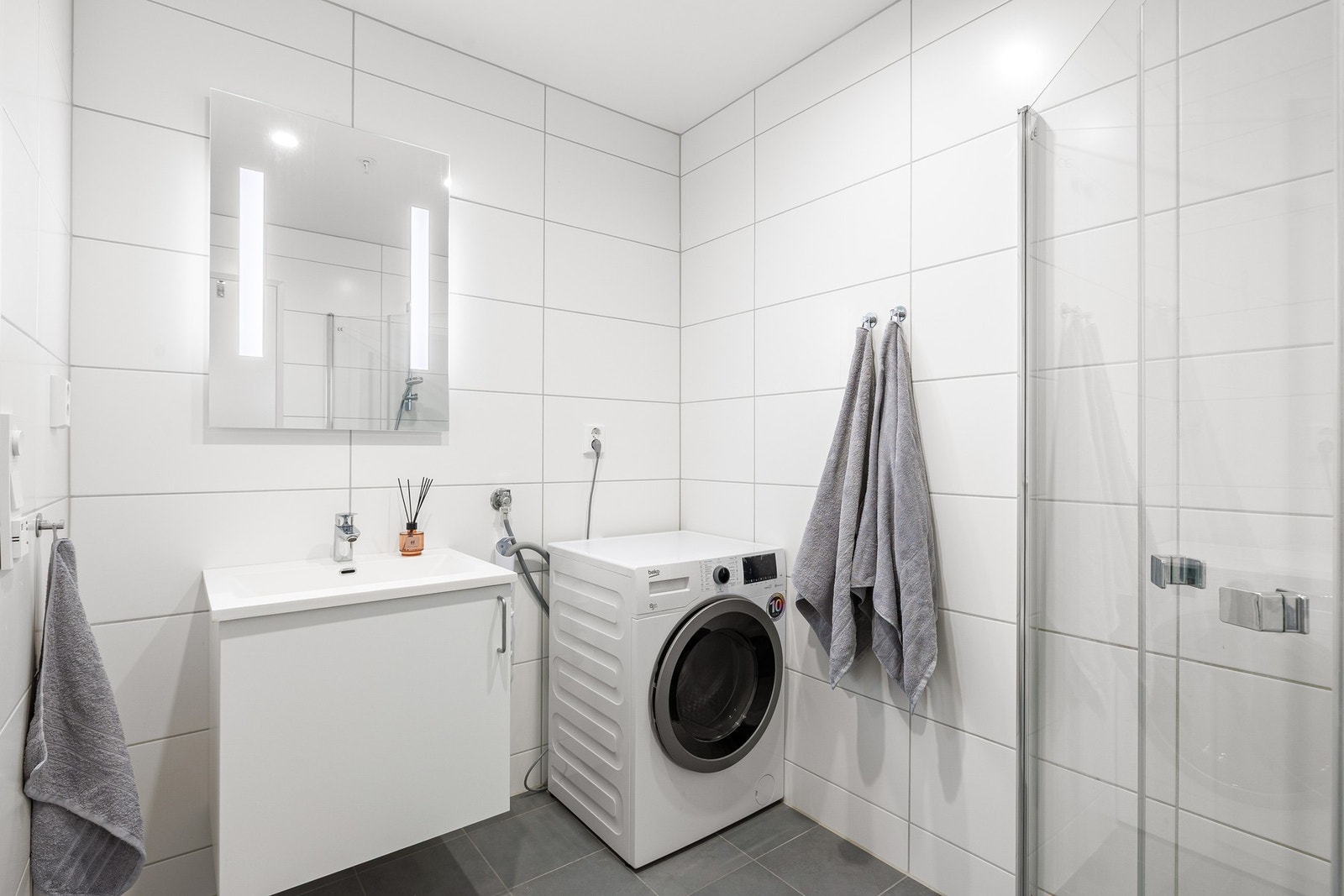 Badet inneholder innfellbare og plassbesparende dusjdører i glass, dusjbatteri og garnityr montert på vegg, kran og avløp for vaskemaskin og veggmontert WC. Galleribilde