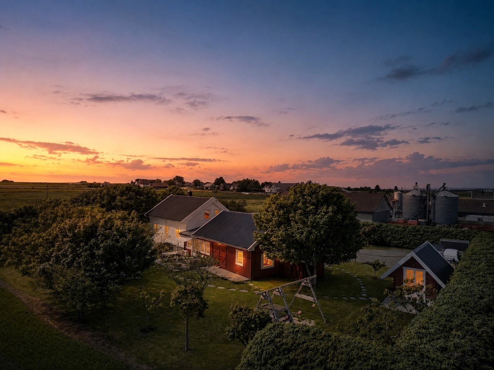Et stemningsfullt kveldslys hviler over et idyllisk gårdstun omgitt av åpne jorder. Solen er i ferd med å gå ned, og himmelen er fylt med et vakkert fargespill. Galleribilde