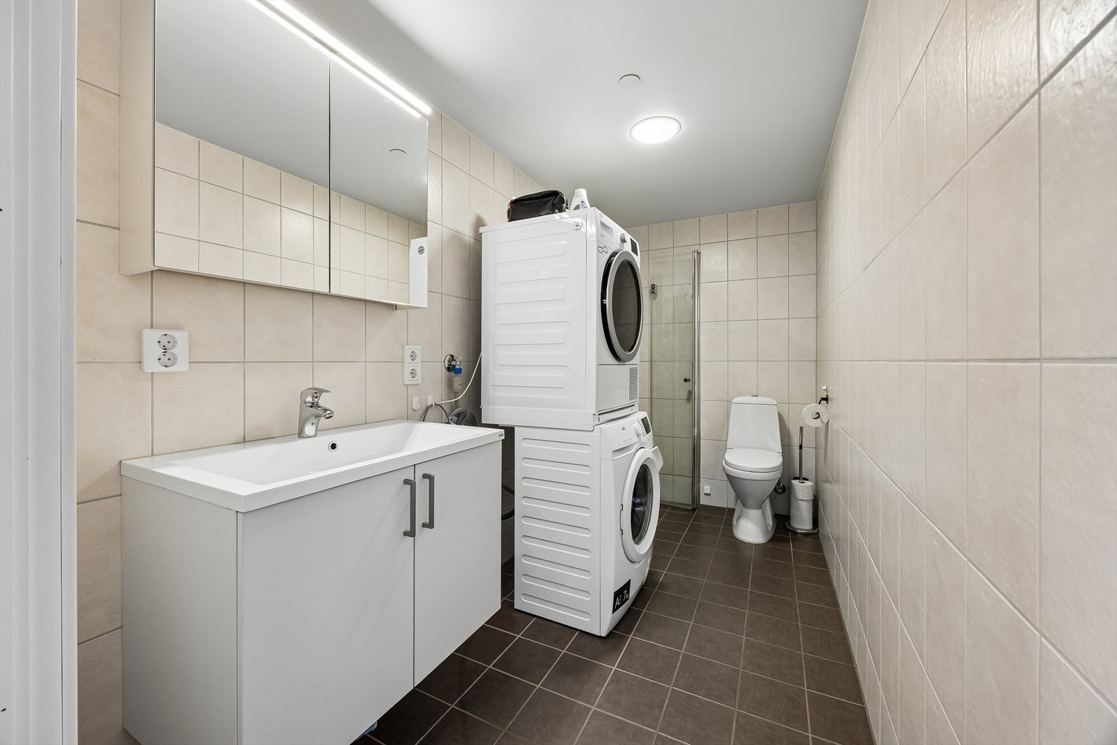 Det helflisede badet har gulvstående toalett og dusjhjørne med innfellbare dører. Innredningen består av heldekkende servant over hvite servantskap og speilskap med belysning i overkant. Galleribilde