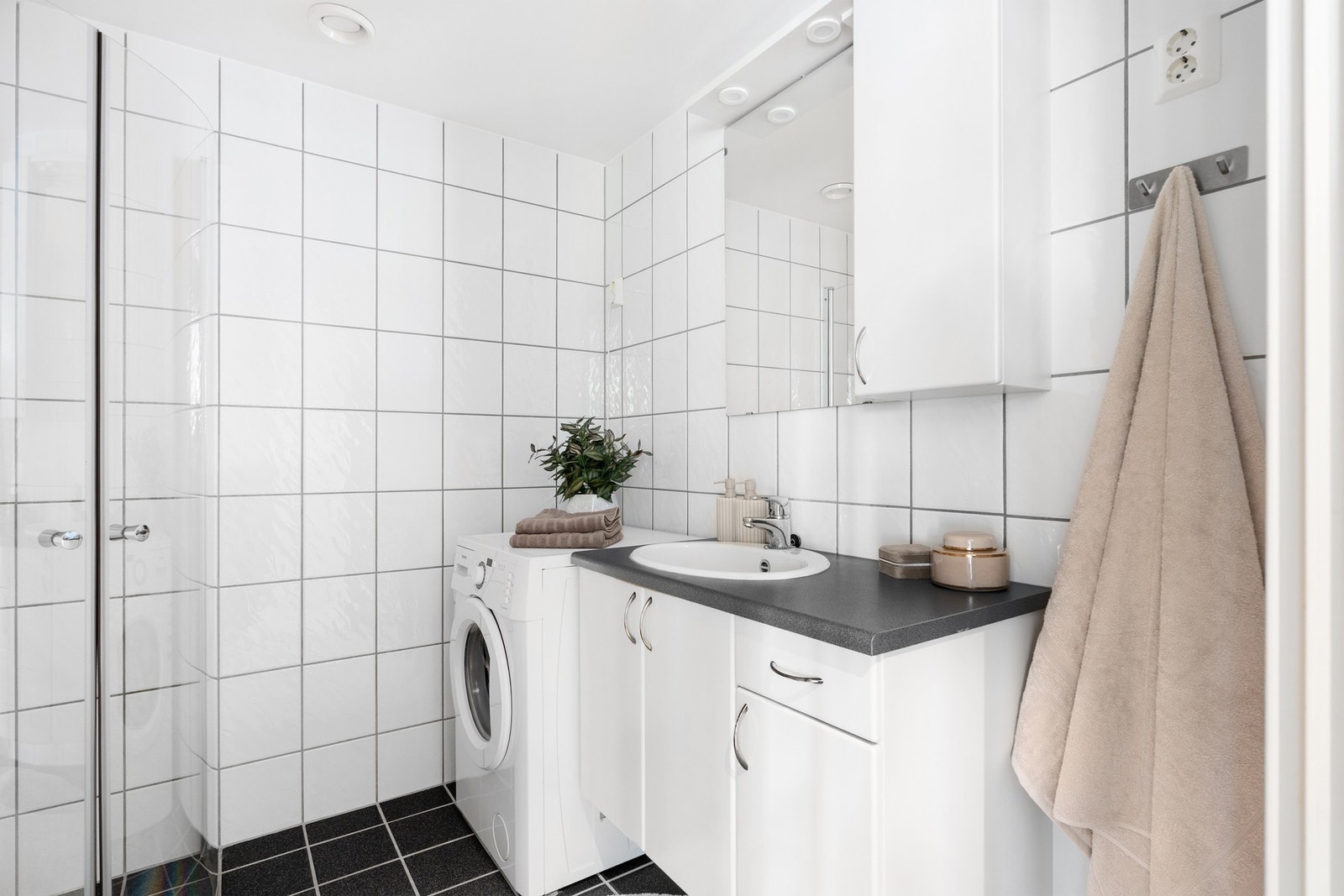 Baderommet inneholder dusjhjørne, kran og avløp for vaskemaskin, gulvmontert WC, servant med underskap, overskap og speil. Galleribilde