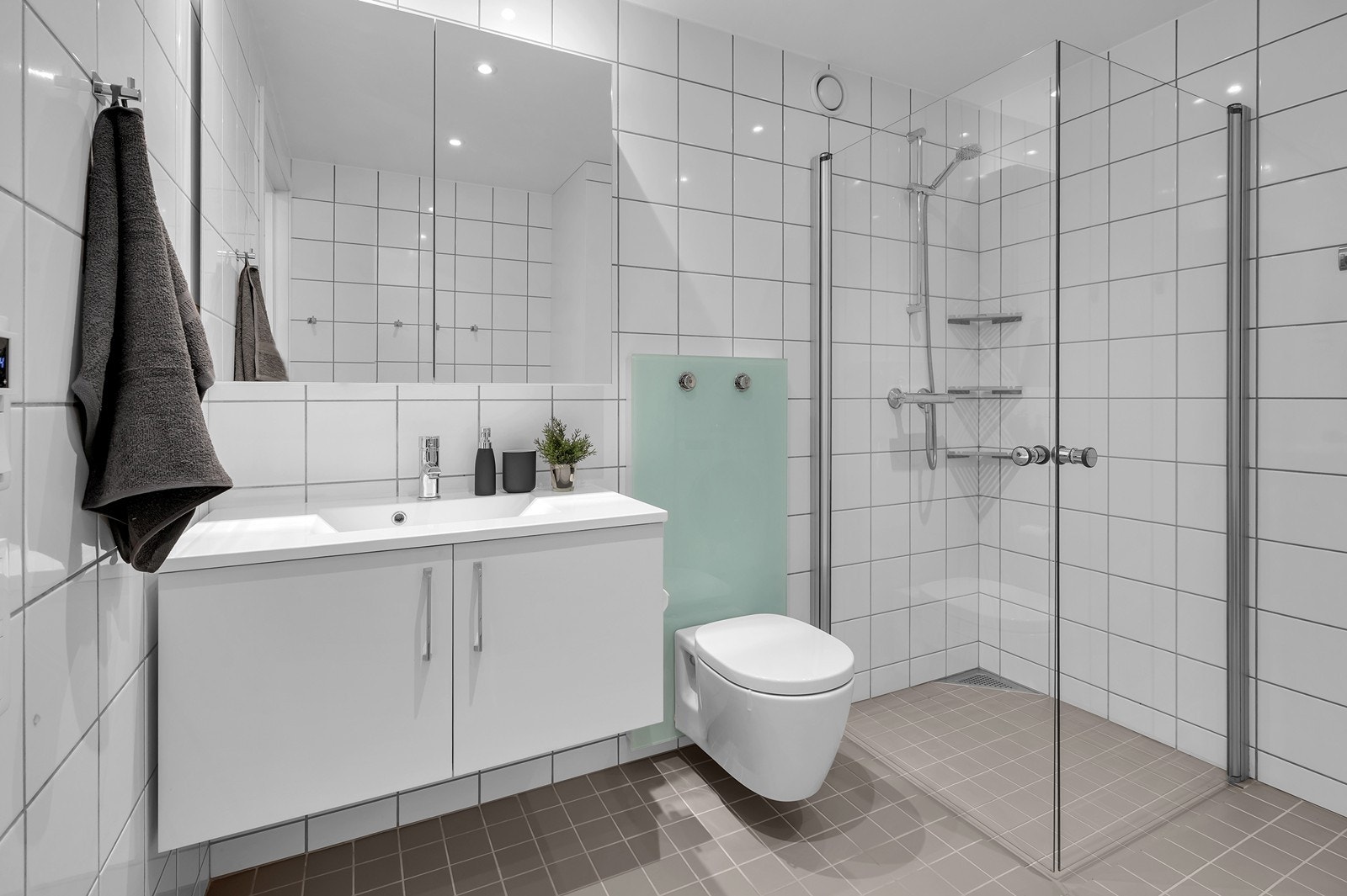 Badet er innredet med flislagte vegger og gulv, malt gipshimling, vegghengt toalett, dusjhjørne, servantinnredning med nedfelt vask og opplegg for vaskemaskin Galleribilde