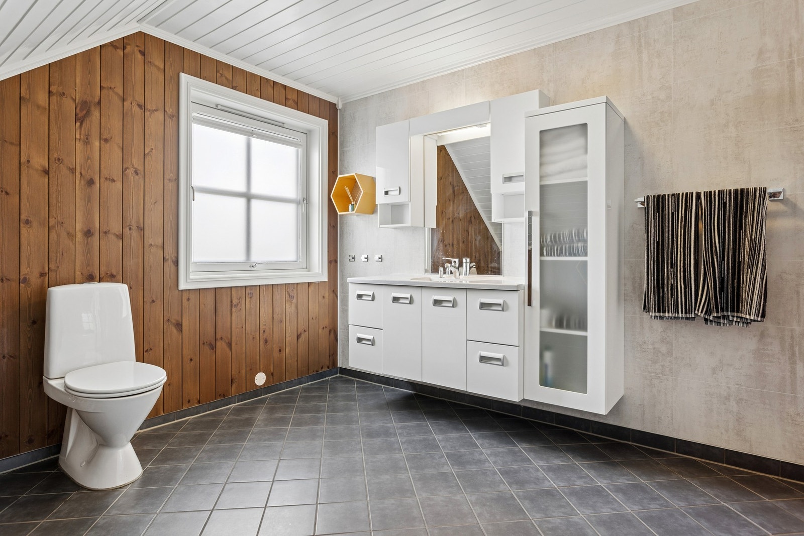 Badet har flislagt gulv, lys baderomsinnredning med rikelig oppbevaringsplass og både badekar og separat toalett. Galleribilde