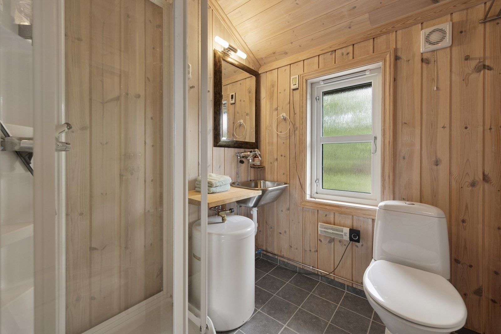 Et praktisk bad med flislagt gulv og varmekabler. Veggene er kledd med panel, og badet er innredet med dusjkabinett, vaskekum og gulvmontert toalett. Galleribilde