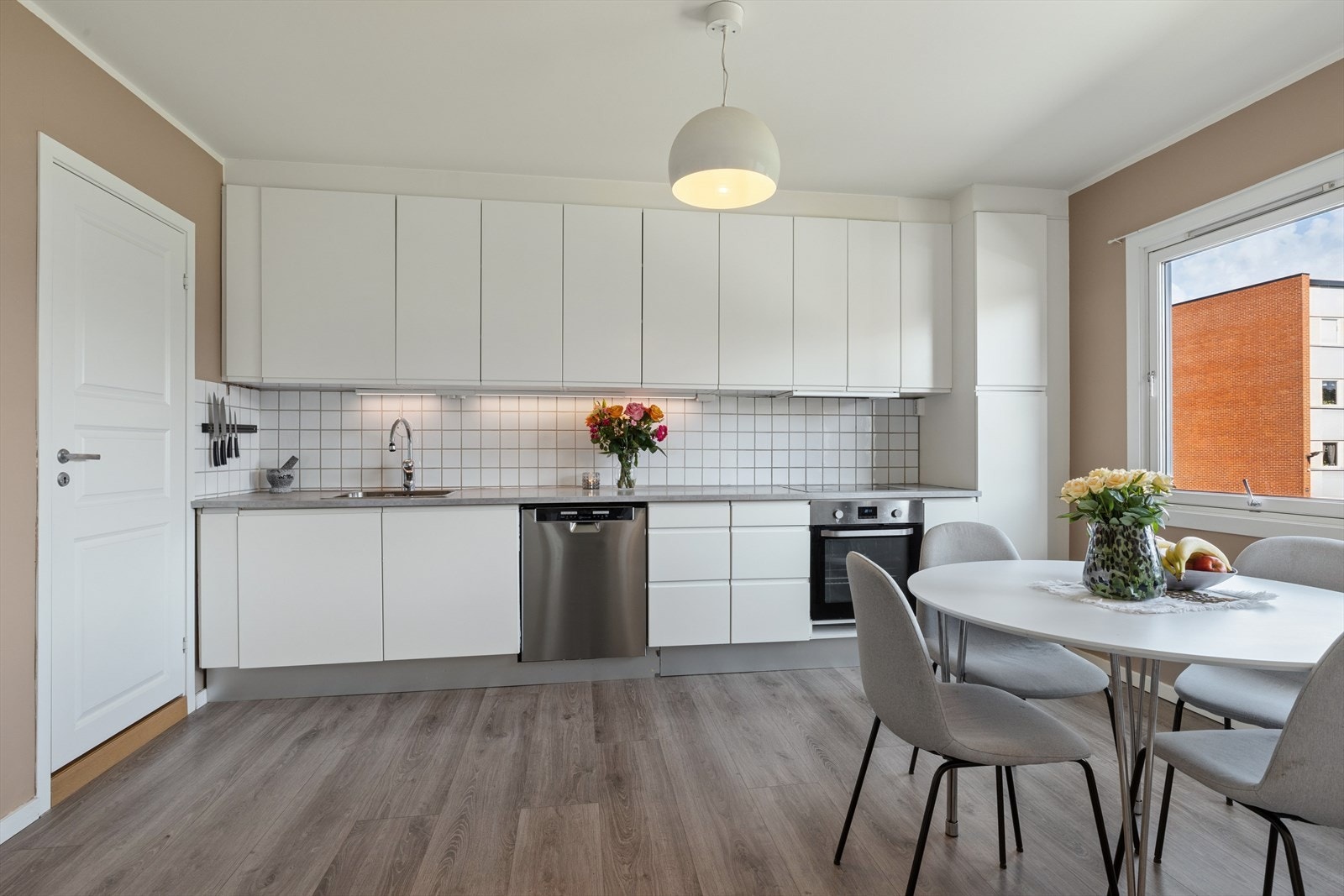Stilrent kjøkken med hvite, slette fronter og fliser over benk. Grå benkeplate gir et moderne preg, og den romslige løsningen gir god plass til en spisegruppe. Galleribilde