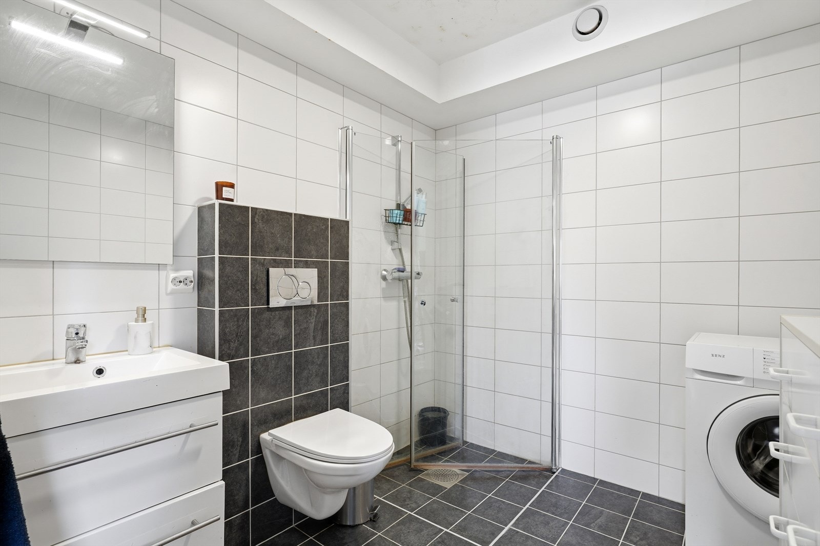 Baderom innredet med dusjsone, toalett, opplegg til vaskemaskin og servant m/skuffer. Galleribilde
