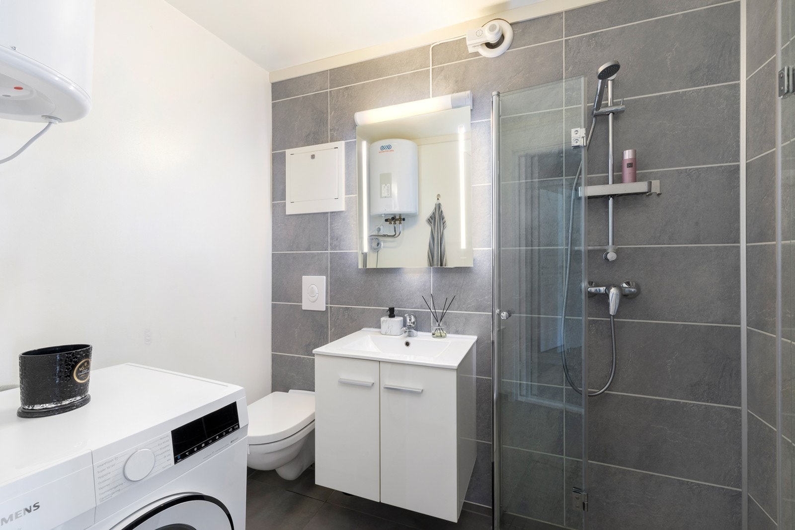 Badet er delvis flislagt med varme i gulvet. Det inneholder en dusjnisje med innfellbare dusjdører, toalett, baderomsinnredning med speilskap over, samt plass og opplegg for vaskemaskin Galleribilde