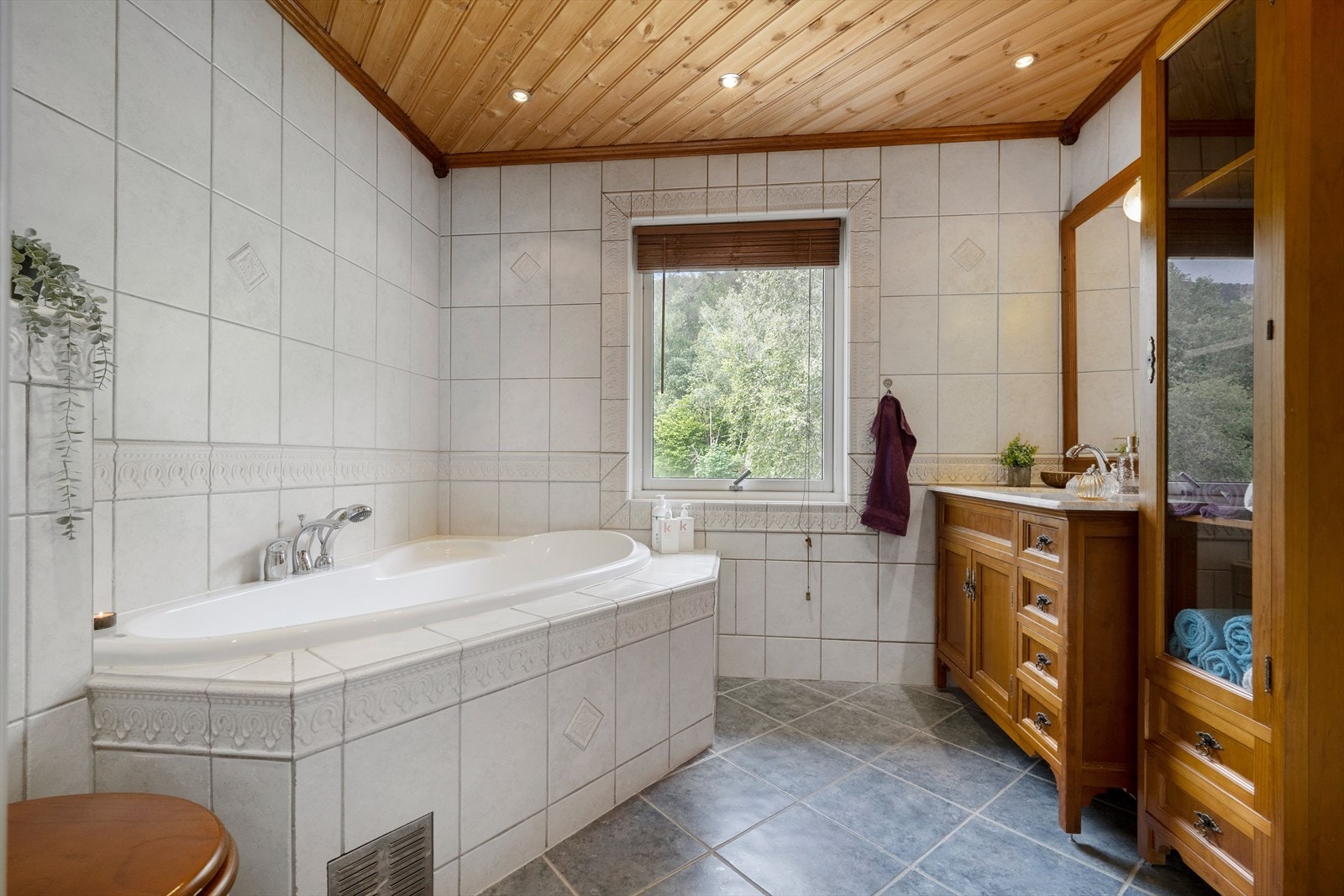 Romslig og funksjonelt bad med flislagte gulv og vegger. Badet er utstyrt med innfliset hjørnebadekar og gulvstående toalett, samt en solid baderomsinnredning i treverk med fine oppbevaringsløsninger. Vinduet gir naturlig lysinnslipp Galleribilde
