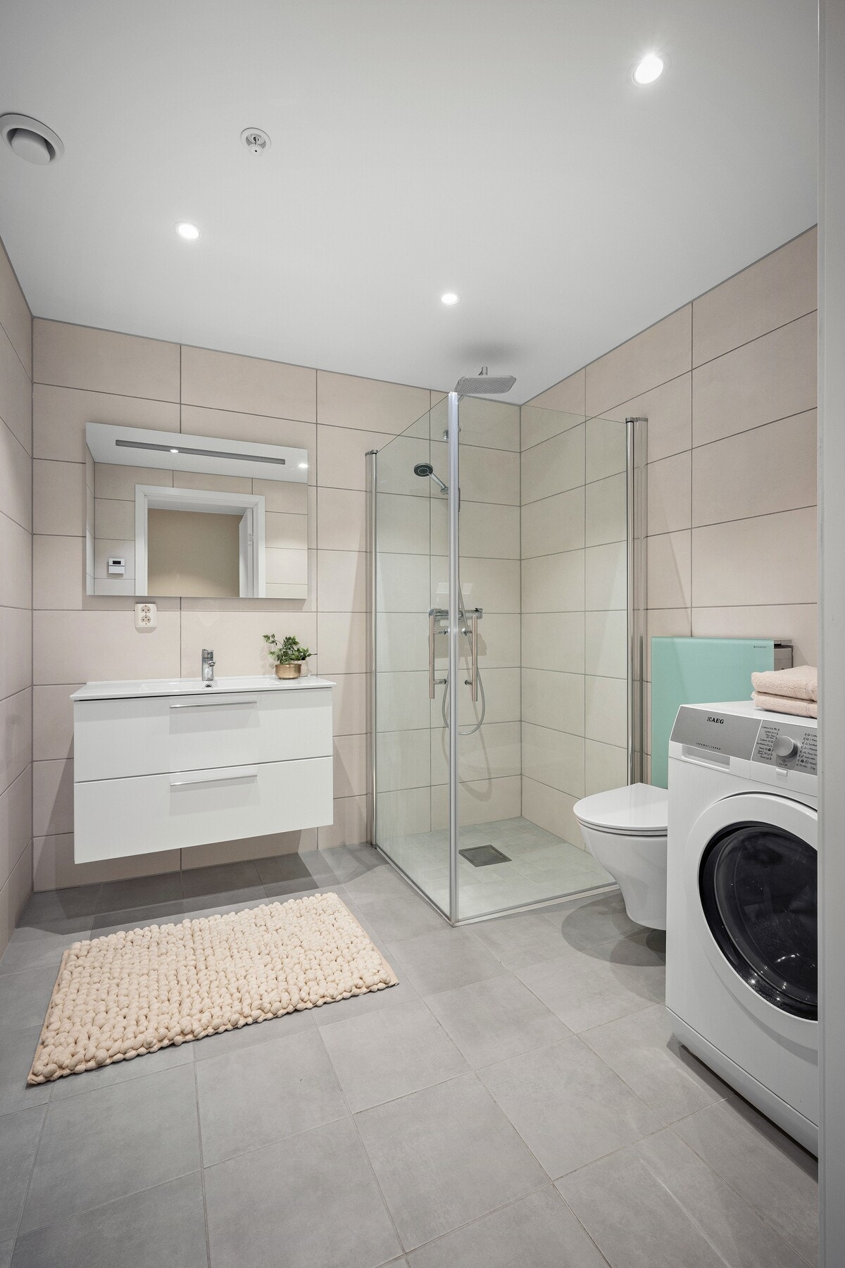 Badet fremstår moderne og delikat, med flislagte overflater, dusjhjørne med glassvegger, vegghengt toalett og stilren baderomsinnredning med god oppbevaringsplass. Rommet har også opplegg for vaskemaskin. Galleribilde