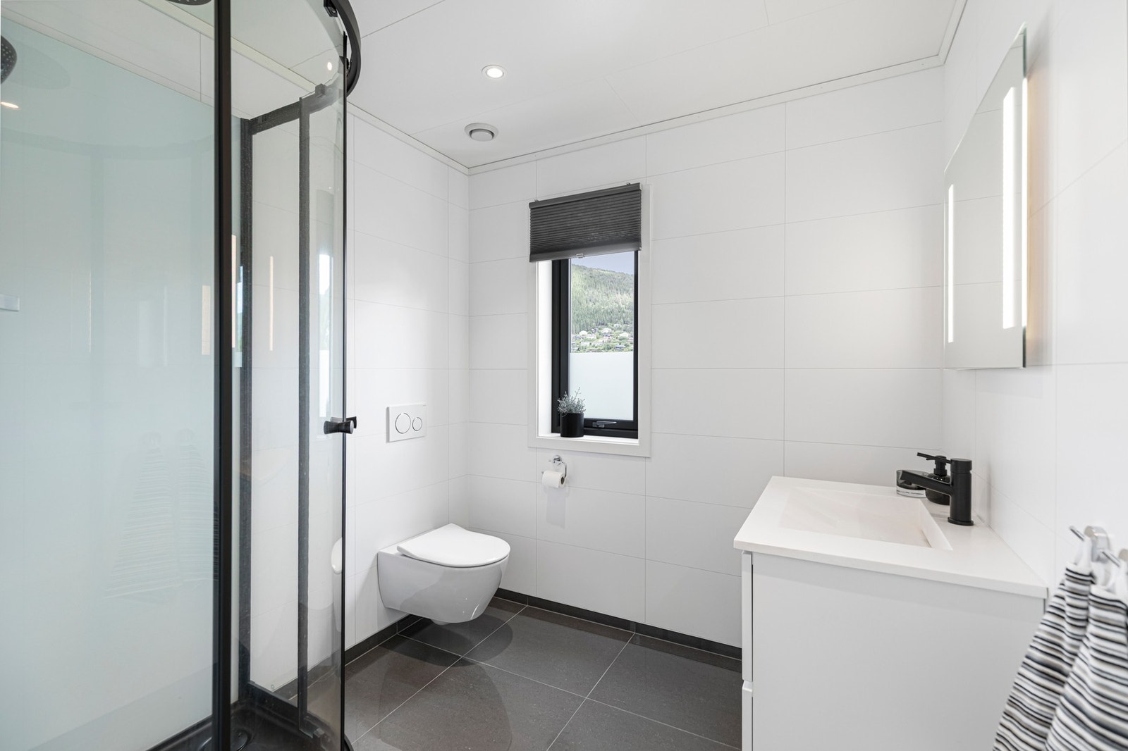 Badet i hovedetasjen er helfliset og er utstyrt med varmekabler i gulv, dusjkabinett, vegghengt toalett og baderomsinnredning med servant, speil og belysning. Galleribilde