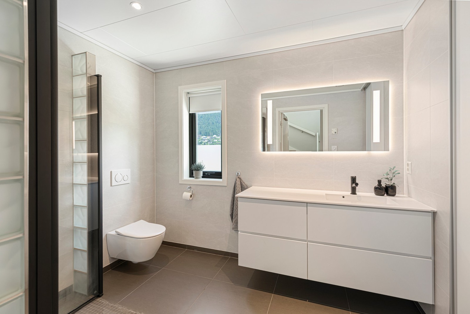Badet i etasjen har flis på gulv og våtromsplater på vegger. Rommet er utstyrt med varmekabler i gulv, baderomsinnredning med servant, dusj med dusjvegger og vegghengt toalett. Galleribilde