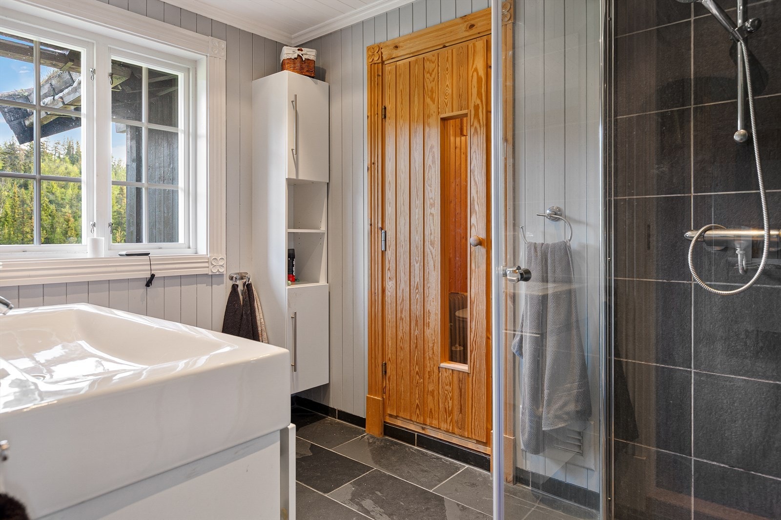 Badet er utstyrt med toalett og dusjhjørne med glassdører og ettgreps dusjarmatur. Baderomsinnredningen består av høyskap og 100 cm servantskap med glatte hvite fronter, servant og ettgreps krummet armatur. Galleribilde
