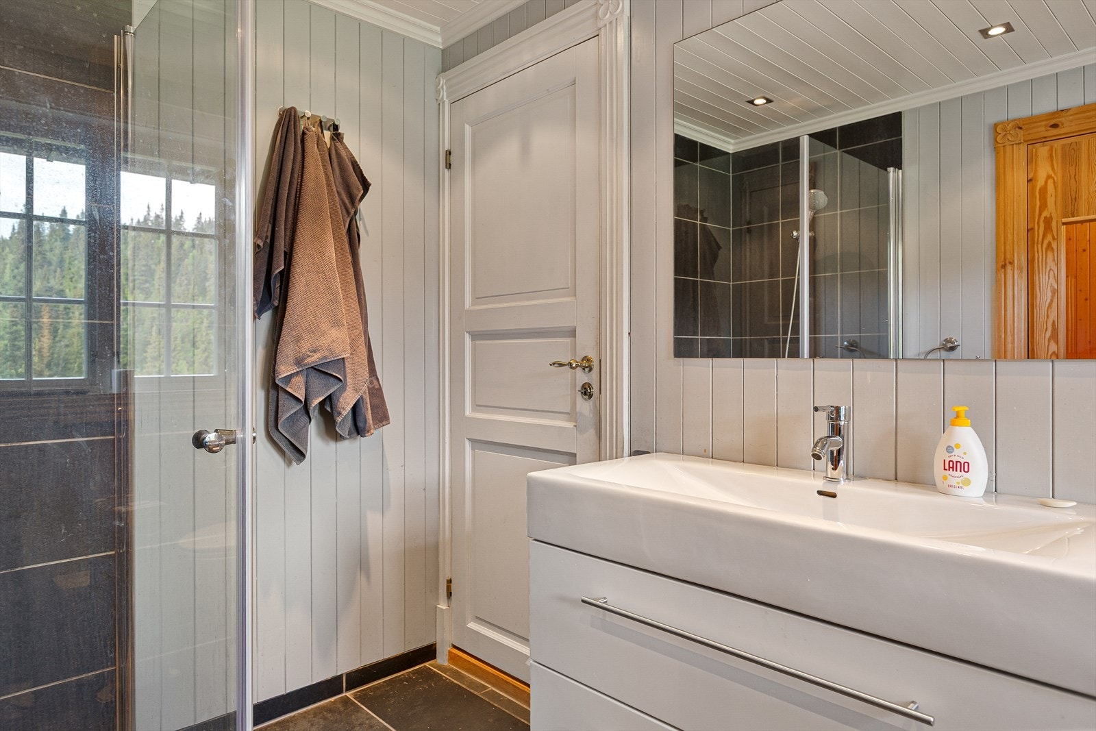 Badet har mørke 30x60 cm fliser på gulv, med 30x30 cm fliser i det nedsenkede dusjområdet og veggene er kledd med mørke 30x30 cm fliser kombinert med malt panel. Galleribilde