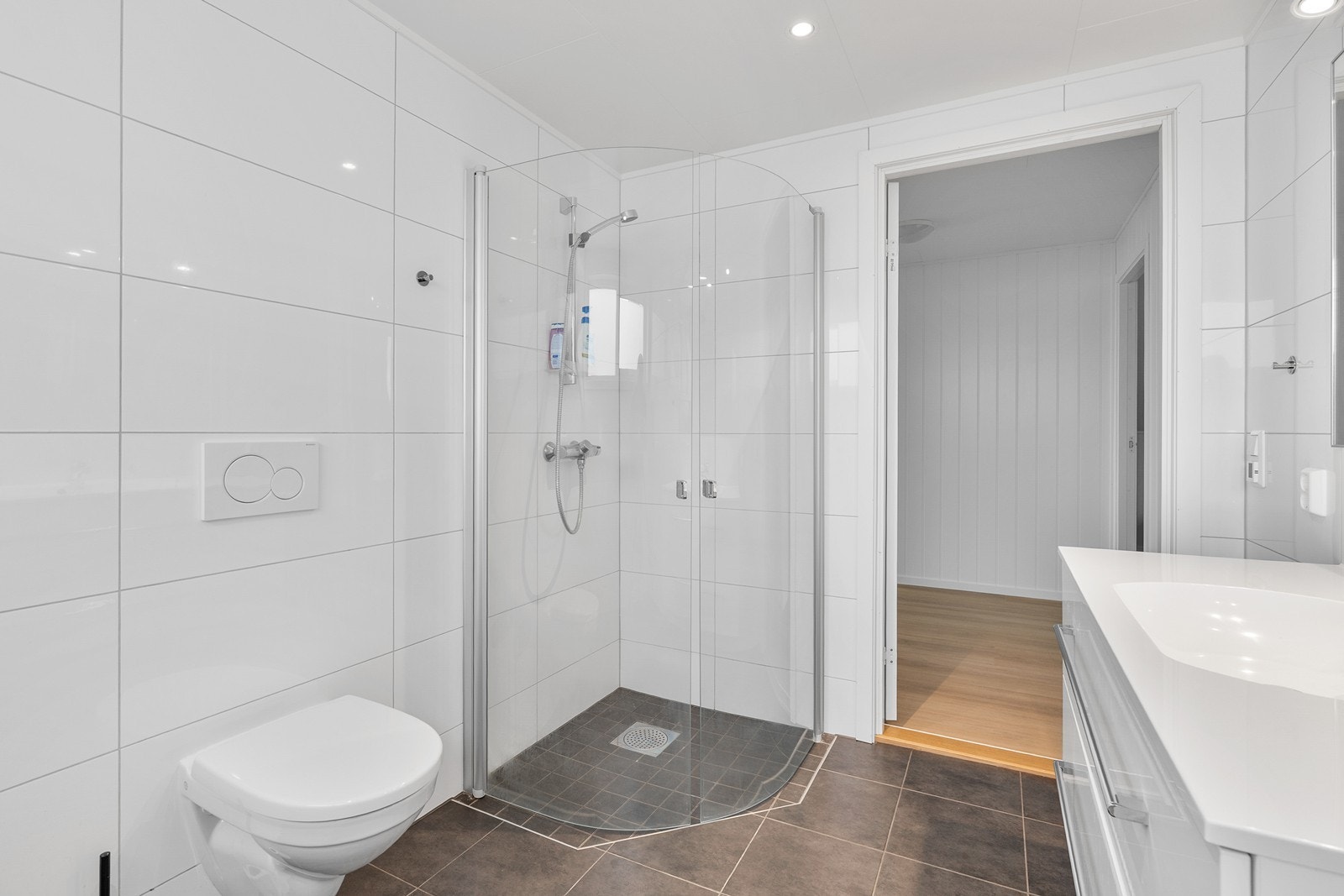 Badet er innredet med vegghengt toalett, dusjhjørne og vask med skuffeinnredning. Galleribilde