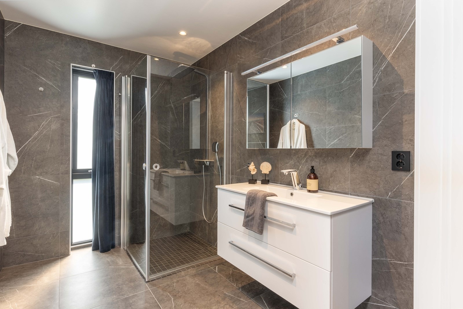 Badet har et moderne og eksklusivt uttrykk, med keramiske fliser på gulv og vegger, og innfelte spotter i taket. Galleribilde