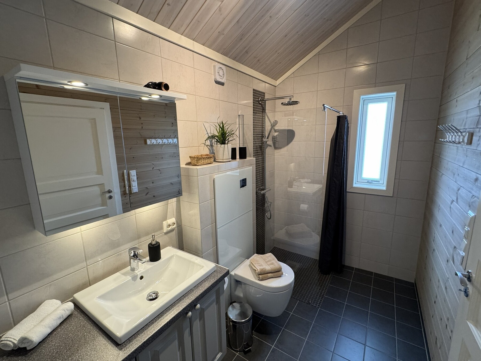 Badet er romslig, og er innredet med mørkere fliser med varmekaber på gulv, og lysere fliser på tre av veggene. Den siste veggen har liggende, beiset panel. Galleribilde