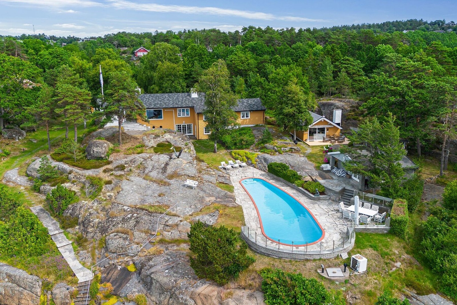 Dronebilde som viser eiendommens unike beliggenhet ved sjøen, med egen brygge, basseng og flere bygninger. Galleribilde