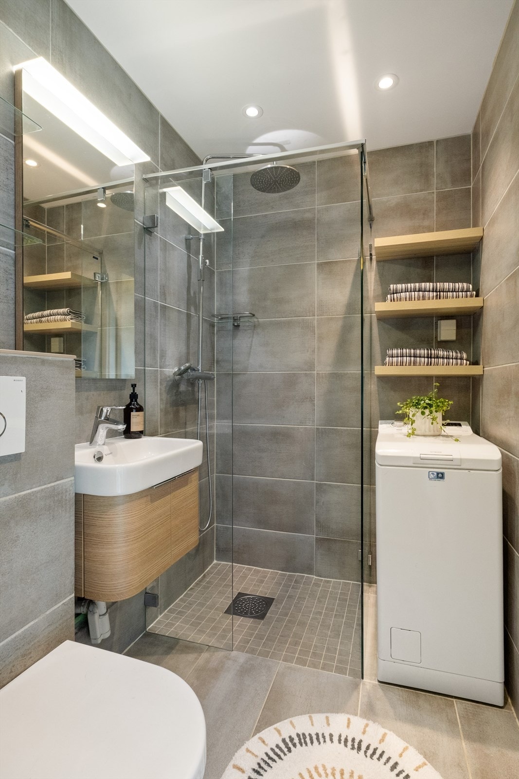 Moderne, flislagt baderom med varme i gulv og downlights i himling. Badet er innredet med nedfelt servant med oppbevaring i underliggende skuffer, veggfestet wc og dusj på gulv med dusjvegger. Galleribilde