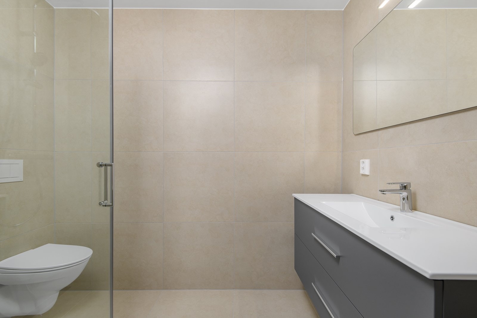 Det største badet er flislagt med beige fliser på gulv og vegg, med grå baderomsinnredning. Galleribilde