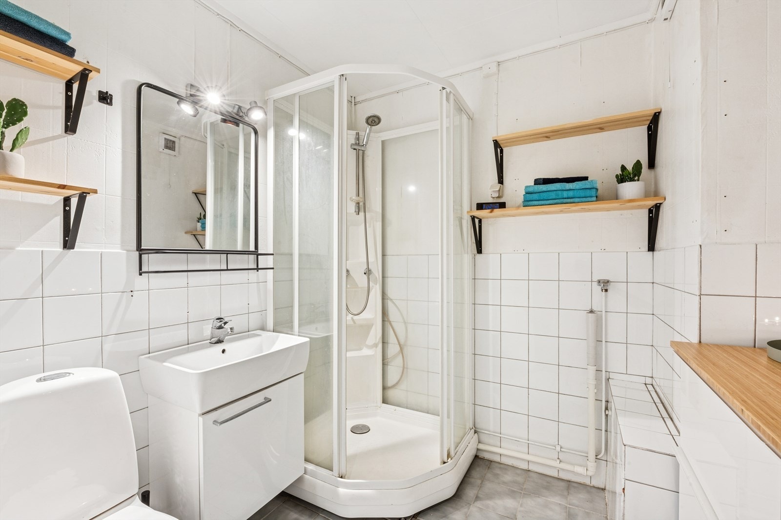 Badet er utstyrt med dusjkabinett, opplegg for vaskemaskin, gulvmontert toalett og servant. Gulvet har fliser og det er flis og tapet på veggene. Galleribilde