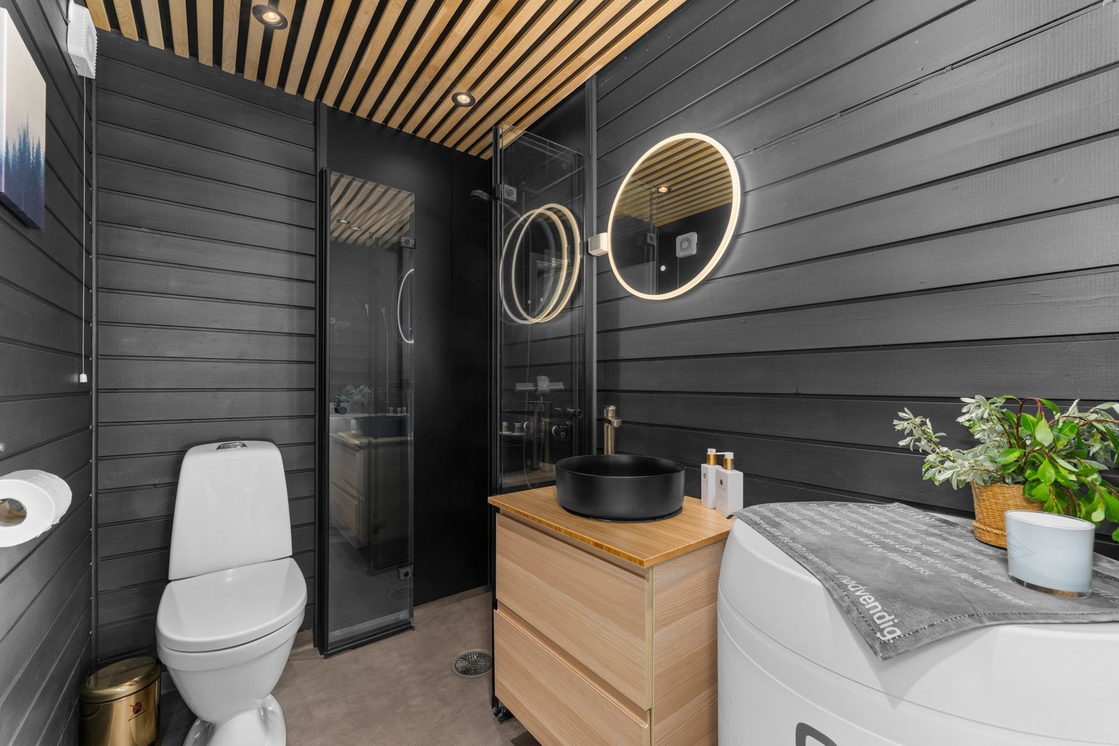 Badet har et moderne og helhetlig uttrykk med mørke vegger, panel og baderomsplater i dusjsonen, spilepanel i himlingen og vinylbelegg på gulvet med elektriske varmekabler. Galleribilde