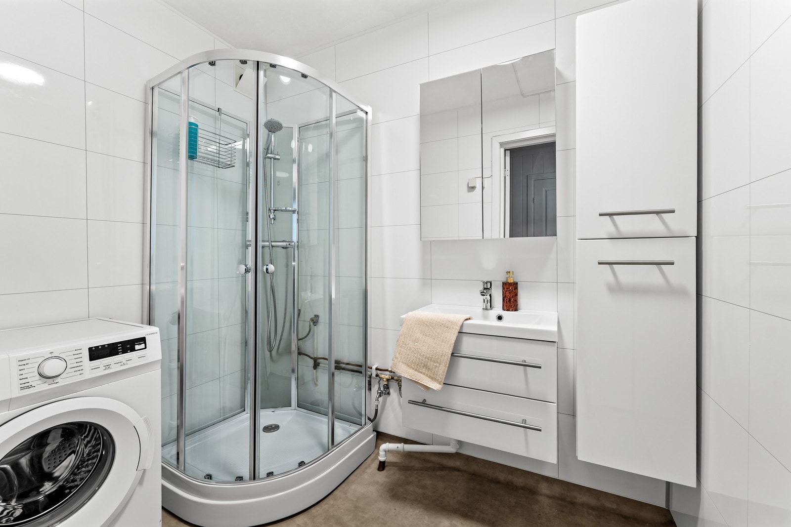 Badet har belegg på gulv og malte, glatte flater i himlingen. Rommet er innredet med en baderomsinnredning med glatte fronter, heldekkende servant med ettgreps blandebatteri, speil med belysning og dusjkabinett. Galleribilde