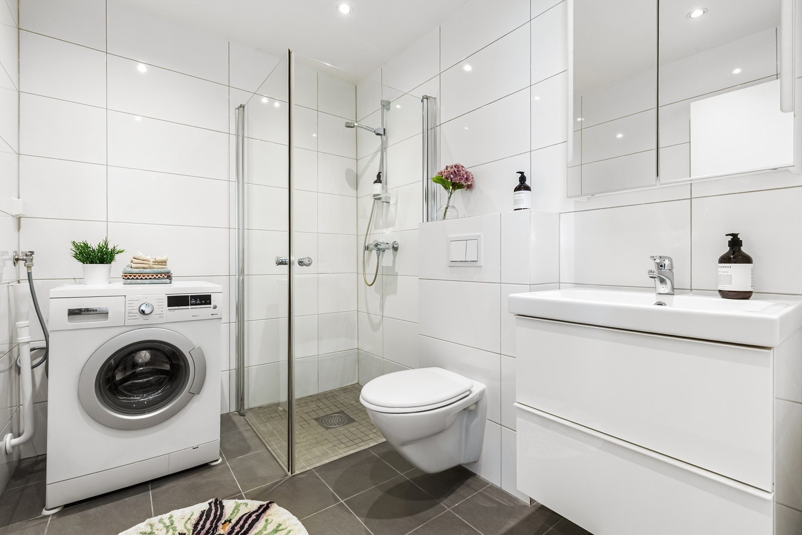 Romslig bad med dusjhjørne, vegghengt wc, servant med skuffer og speilskap. Badet har opplegg for vaskemaskin. Galleribilde