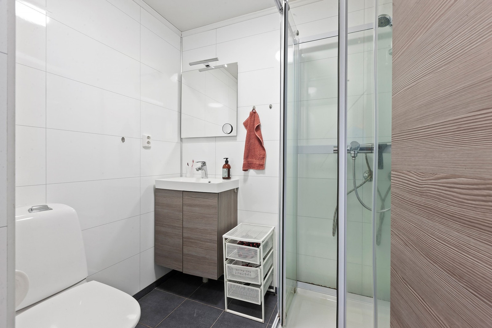 Badet er innredet med mørk baderomsinnredning med nedfelt servant og underskap, speil med belysning over, dusjkabinett og toalett. Praktisk vaskerom ligger i tilstøtende rom. Galleribilde