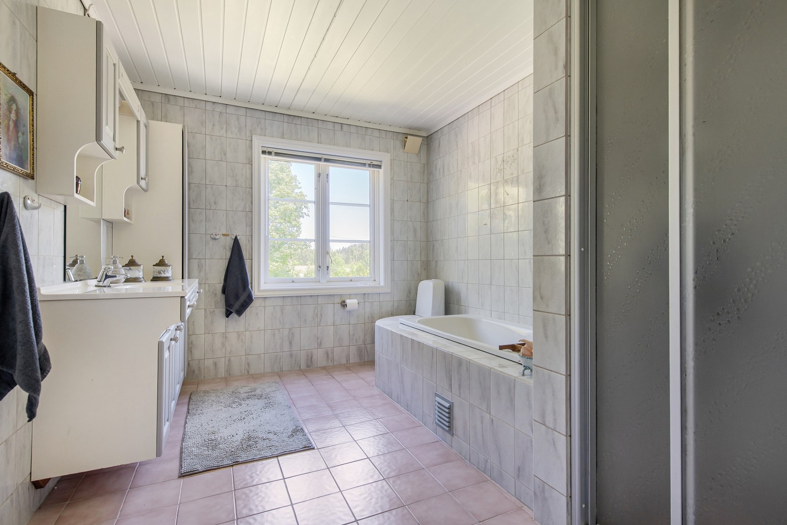 Badet er innredet med grå vegg- og gulvfliser, og har både et dusjhjørne med skyvedører og et innfliset badekar med dusjløsning. Galleribilde