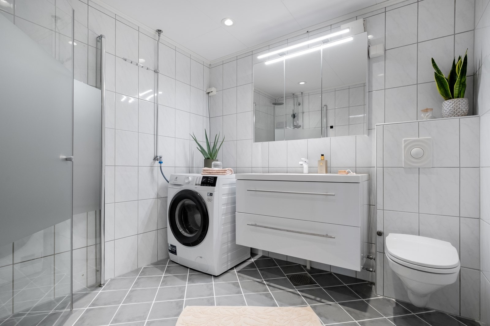 Badet er innredet med nedfelt servant, veggmontert toalett, dusjvegger og opplegg for vaskemaskin. Galleribilde
