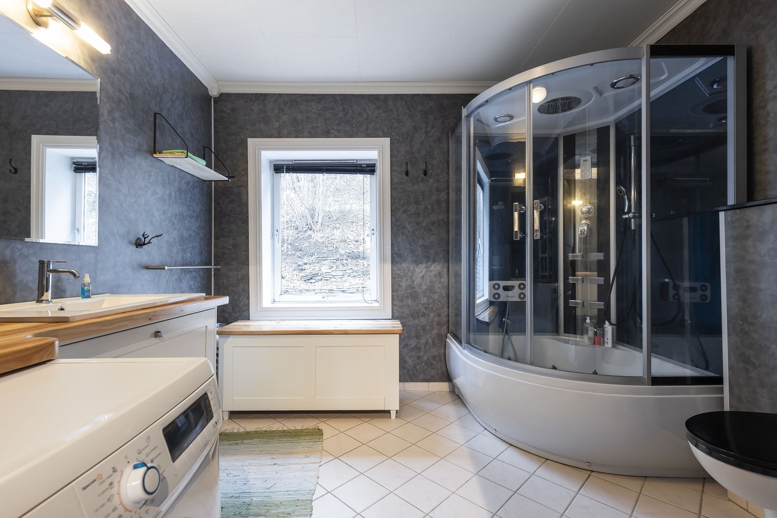 Badet er utstyrt med vegghengt toalett, stort dusjkabinett/badekar, servant med tilhørende innredning og opplegg for vaskemaskin. Galleribilde