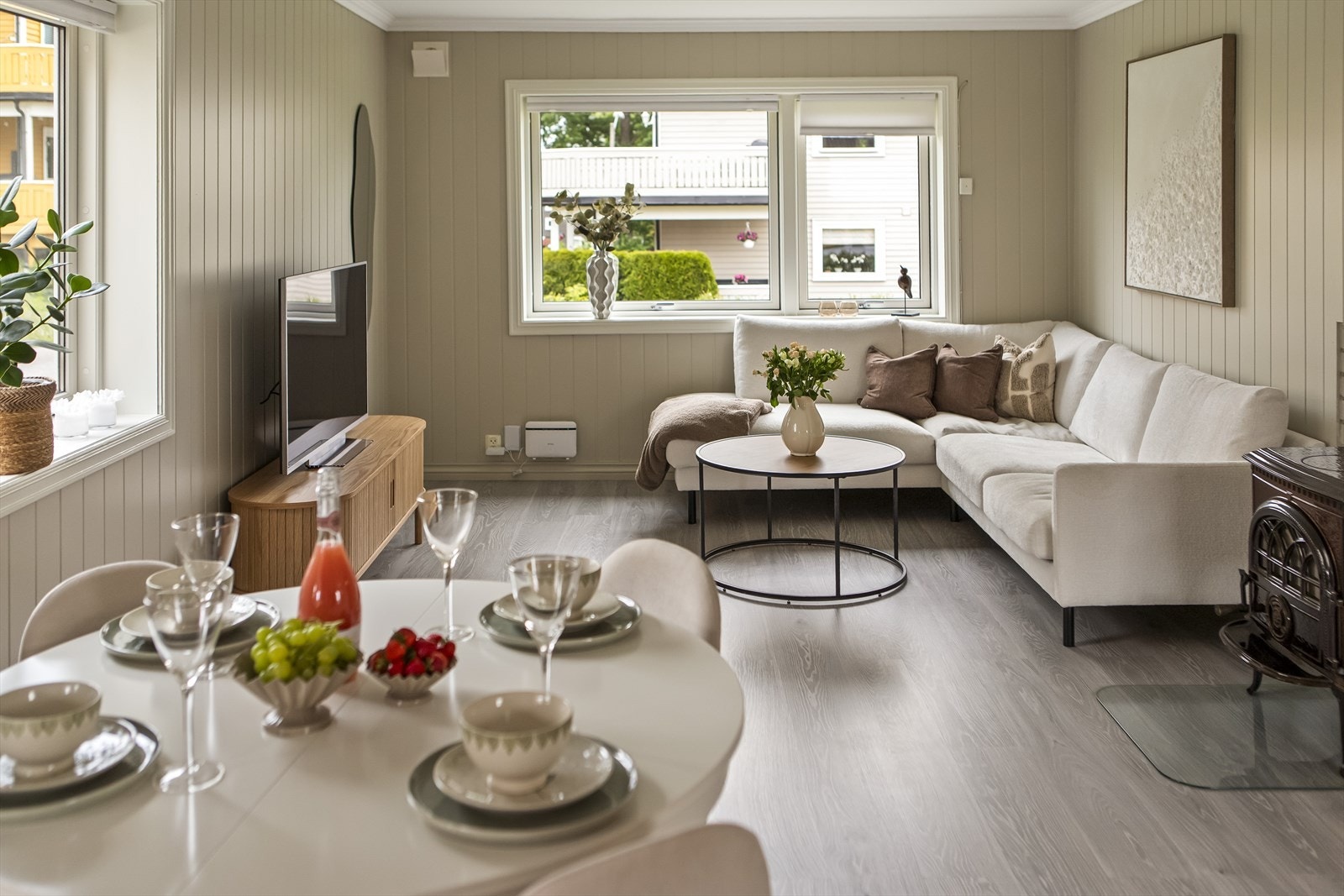 Stuen fremstår som et lyst og luftig oppholdsrom med pen, moderne laminat på gulvet og vegger malt i en behagelig og lys beige farge på stilig panel. Galleribilde