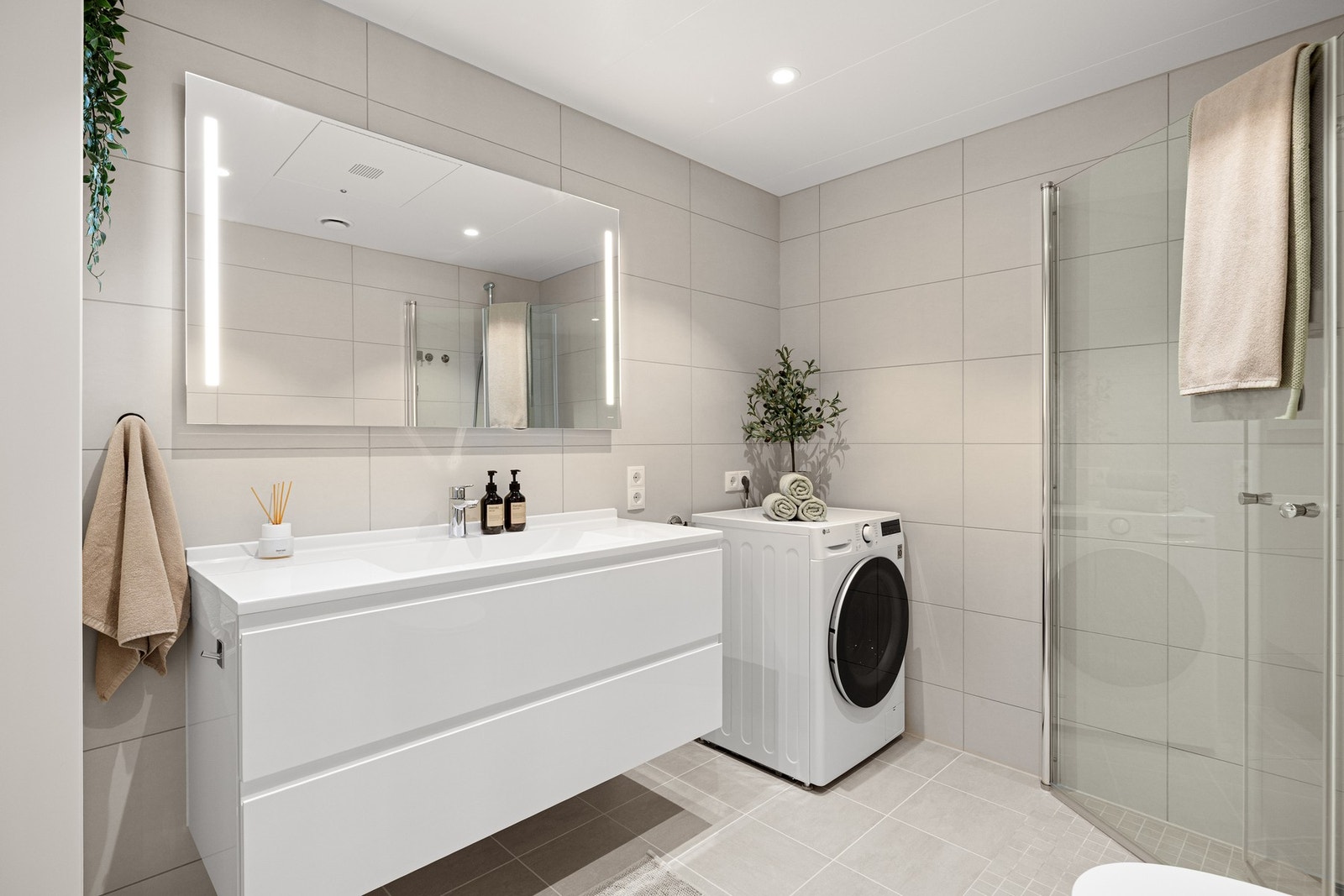 Det lyse og pene badet inneholder innfellbare og plassbesparende dusjdører i glass, dusjbatteri og garnityr montert på vegg, kran og avløp for vaskemaskin og veggmontert WC. Galleribilde