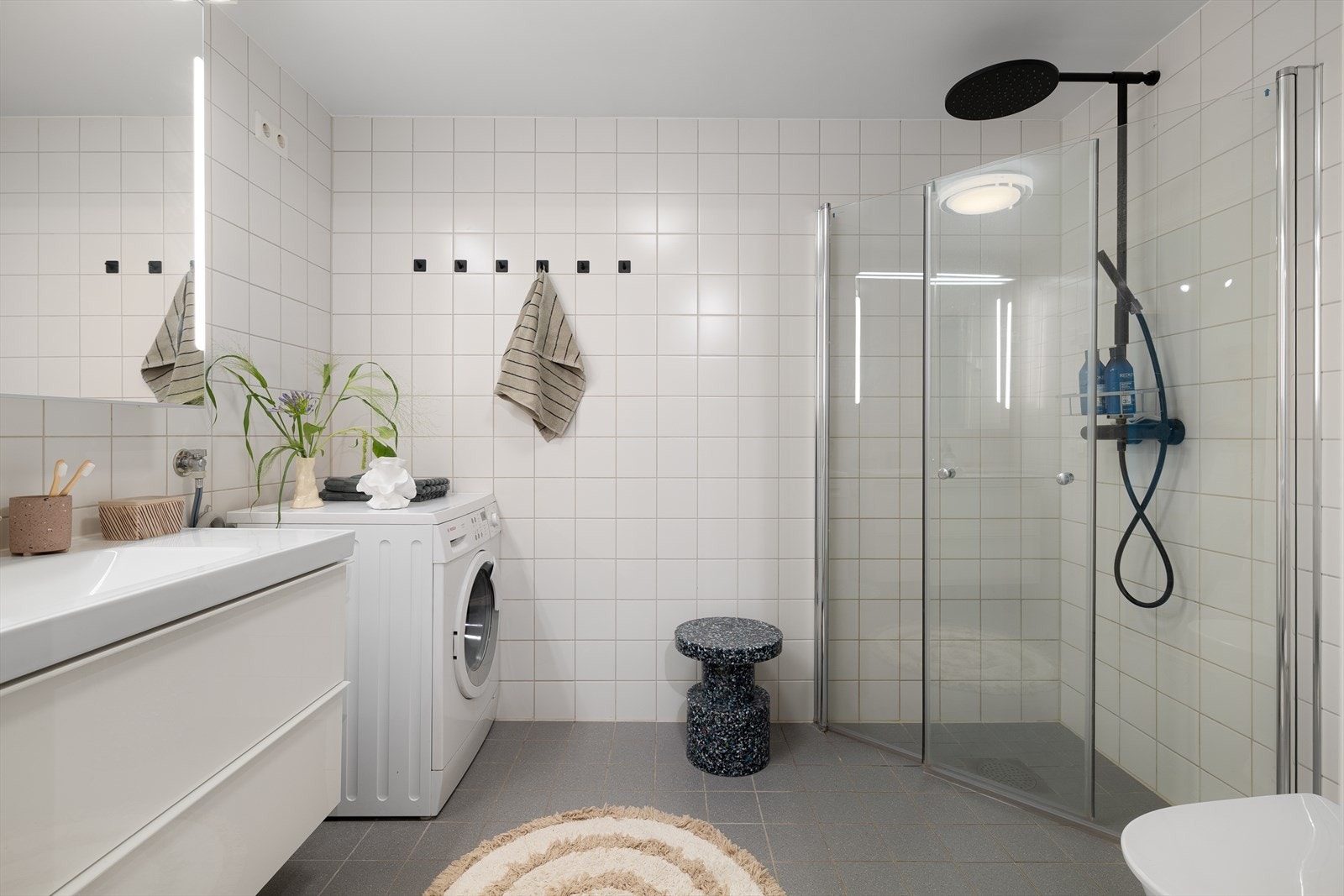 Pent flislagt baderom av behagelig størrelse innredet med klosett, dusjhjørne, servant med innredning og opplegg for vaskemaskin. Galleribilde