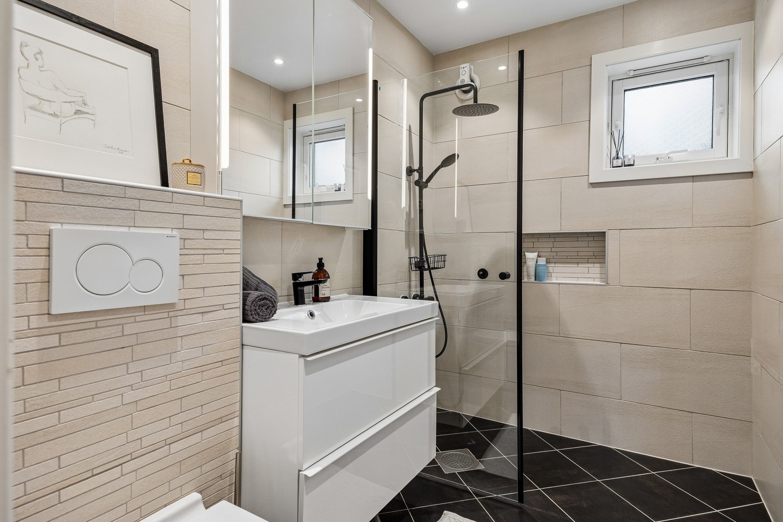Badet holder en moderne standard med flislagte gulv og vegger, malt himling med innfelte downlights og en stilren innredning med servantskap, vegghengt toalett og dusjhjørne. Galleribilde