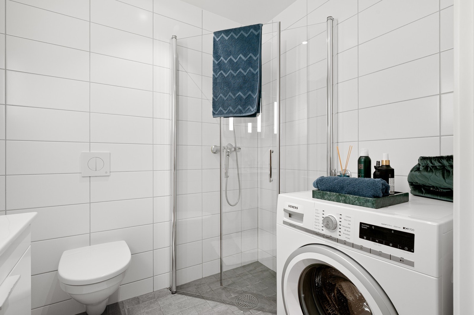Badet inneholder innfellbare og plassbesparende dusjdører i glass, dusjbatteri og garnityr montert på vegg, kran og avløp for vaskemaskin og veggmontert WC. Galleribilde