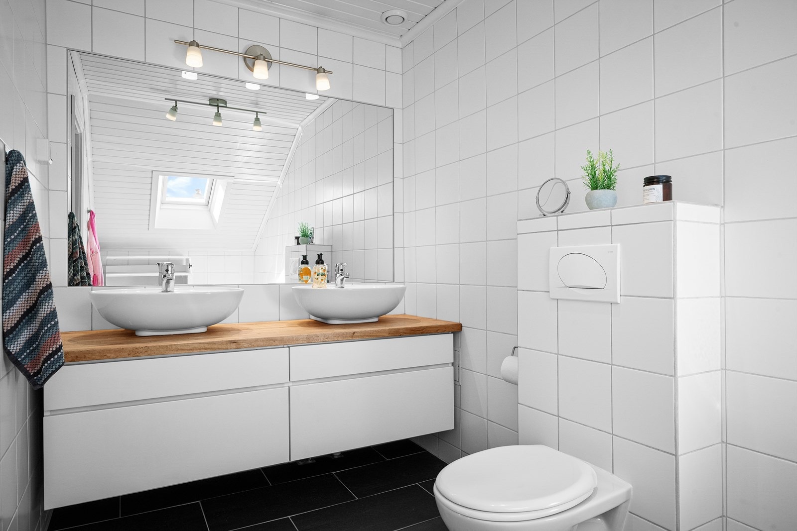 Badet har fliser på gulv med gulvvarme og fliser på vegger. Panel i tak. Seksjon med to vasker, vegghengt toalett med skjult sisterne og innfliset badekar. Galleribilde