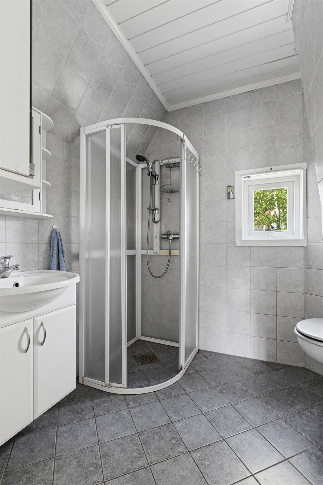 Bad i 2. etasje med fliser på gulv og vegger, panel i himling og varmekabler i gulvet. Badet er innredet med servant med innredning, dusjhjørne og veggmontert toalett, samt ventilator med avtrekk. Galleribilde