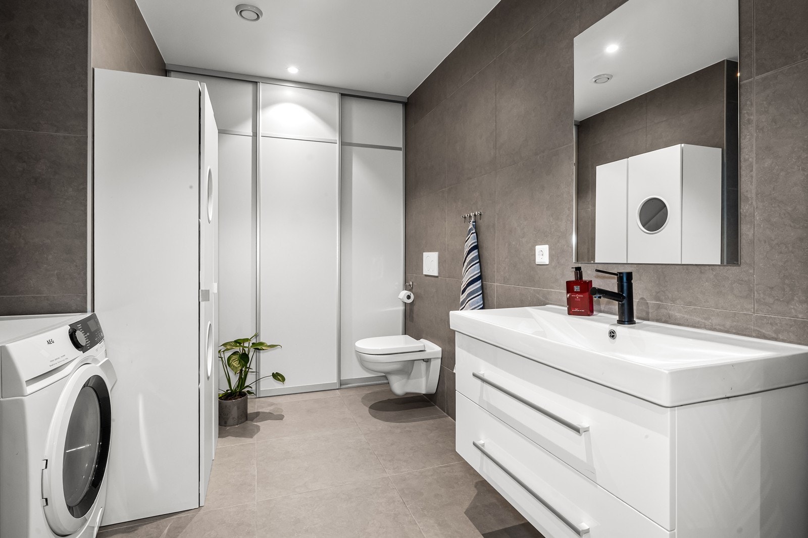 Badet i første etasje er et kombinert vaskerom, med opplegg for vaskemaskin. Dette rommet er også flislagt med moderne 60x60 fliser, med elektriske varmekabler i gulvet. Videre er det servantinnredning med lagringsplass i skuffene, dusj, og vegghengt wc. Galleribilde