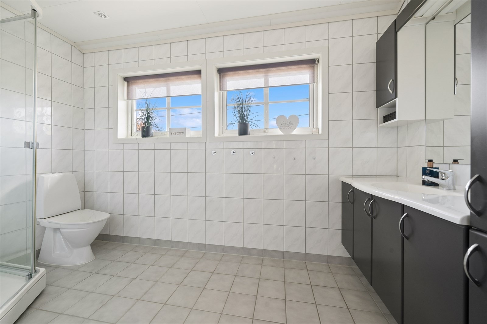 Romslig, flislagt baderom med lyst og tidløst uttrykk. Badet har to vinduer uten innsyn som gir godt med naturlig lys, samt dusjhjørne, toalett og en velutnyttet innredning med god oppbevaringsplass og praktisk benkeplate ved servant. Galleribilde